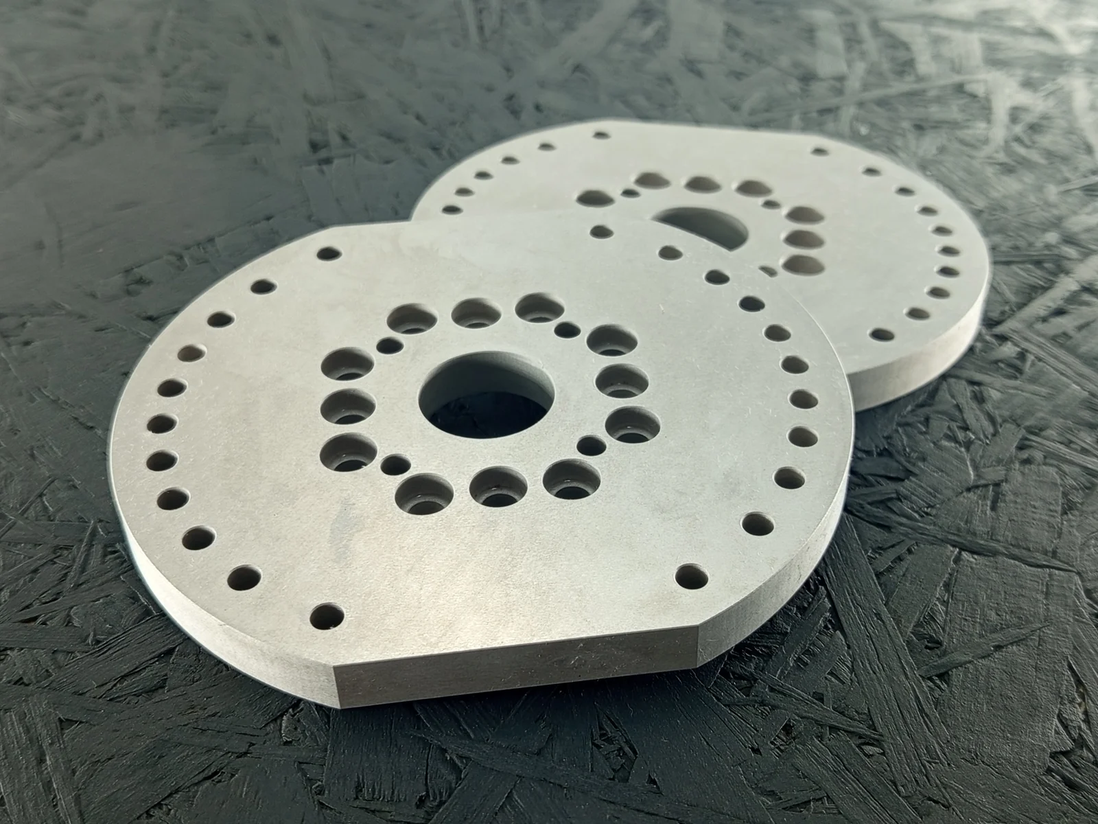 Zwei CNC-gefräste Adapterplatten aus Alu 7075, 10mm dick, mit vielen Bohrungen, auf dunklem Hintergrund.