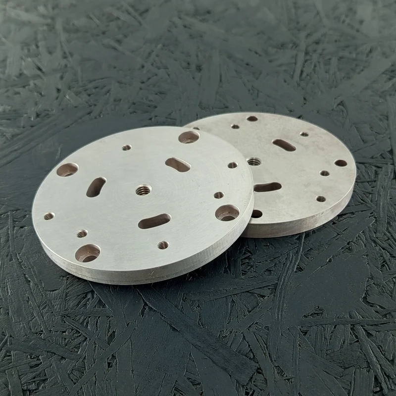 Zwei CNC-gefräste runde Montageplatten aus Alu 7075, 8mm dick, mit Gewinden, Senkungen, Bohrungen und Langlöchern auf dunklem Hintergrund.
